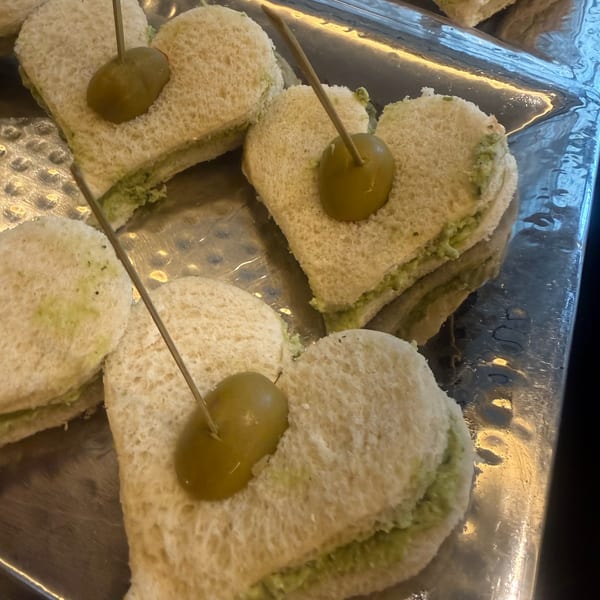 Heart-shaped finger sandwiches with green filling and an olive on a toothpick, on a silver tray.