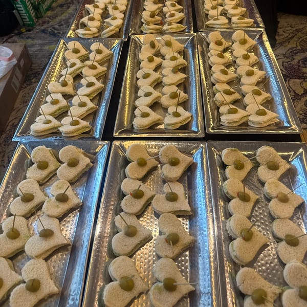 Heart-shaped finger sandwiches with green olive garnishes are arranged on multiple silver trays.