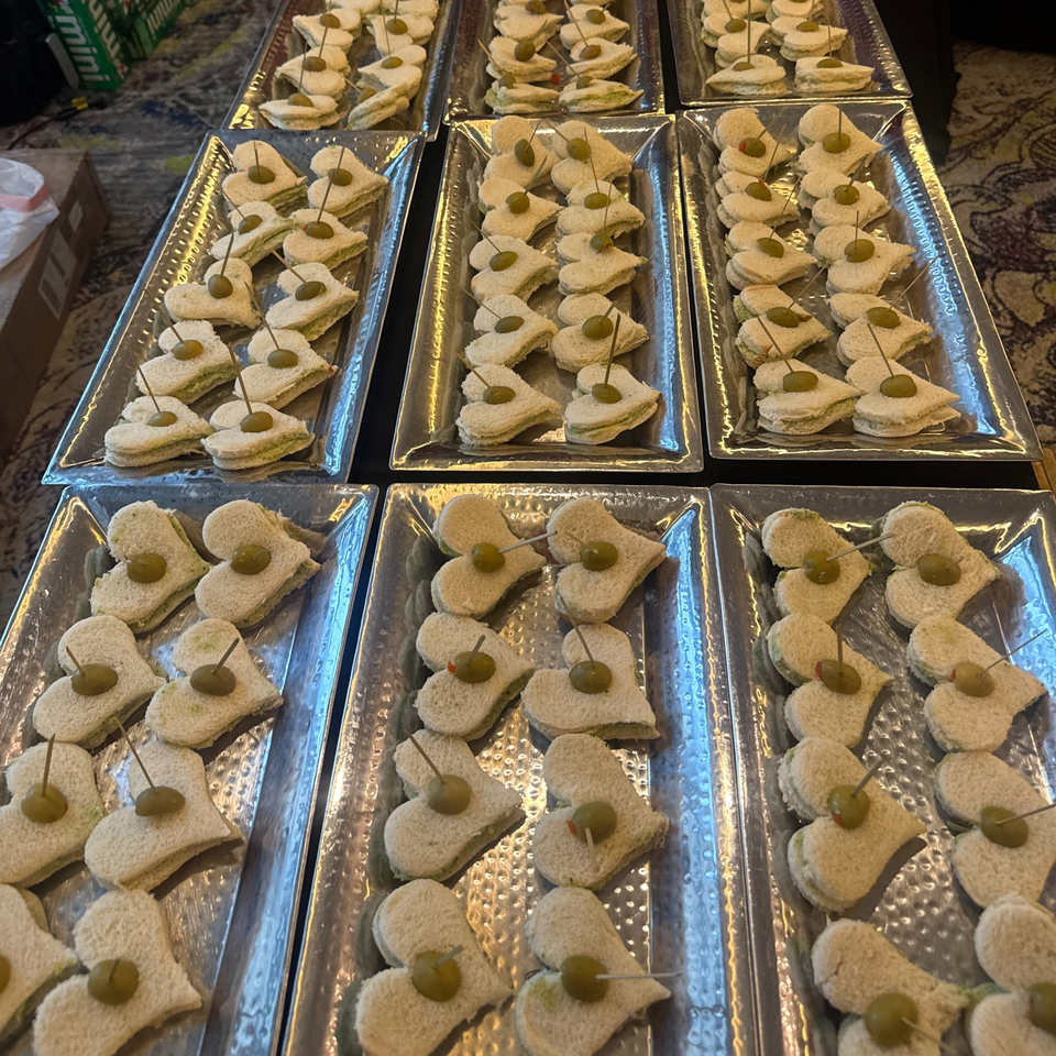 Heart-shaped finger sandwiches with green olive garnishes are arranged on multiple silver trays.