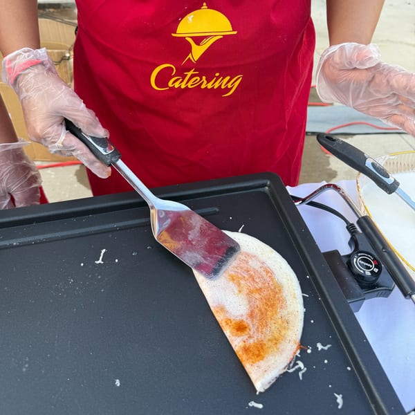 A person in a red catering apron and gloves uses a spatula to flip a dosa on a griddle.