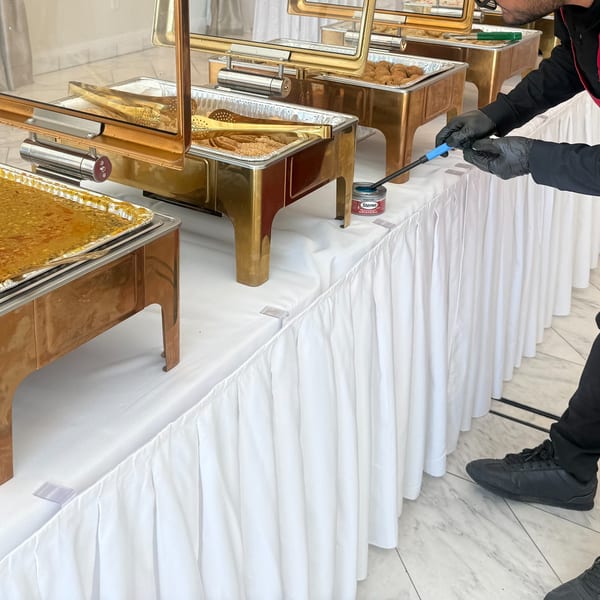 A person in black gloves lights a Sterno can under a gold chafing dish filled with food.