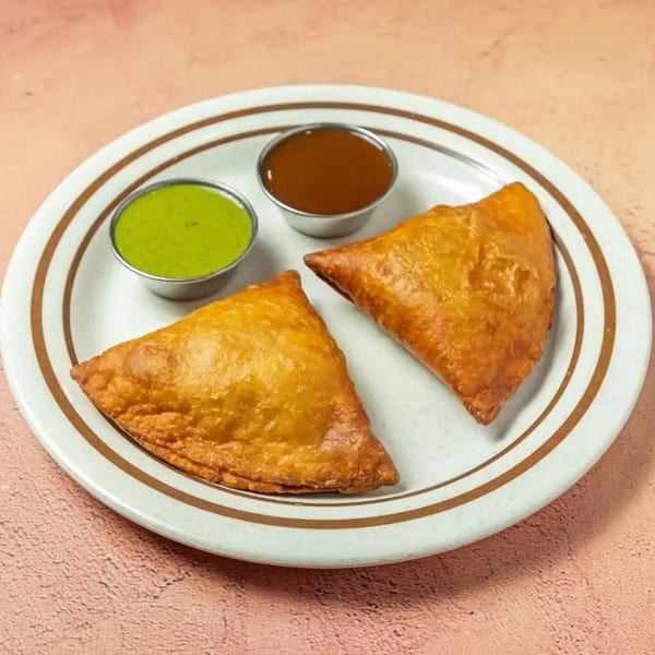 Two golden-brown samosas and two small bowls of chutney on a white plate with brown stripes.