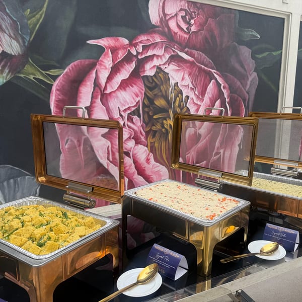 Buffet line with Gujarati Pulao and Dhokla in chafing dishes, against a floral wall mural.