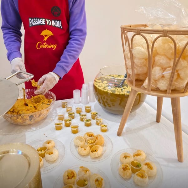 A person in a red Passage To India Catering apron prepares pani puri at a buffet table.