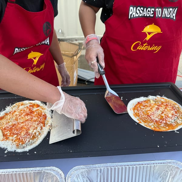 Two chefs in red Passage to India Catering aprons prepare dosas on a griddle.