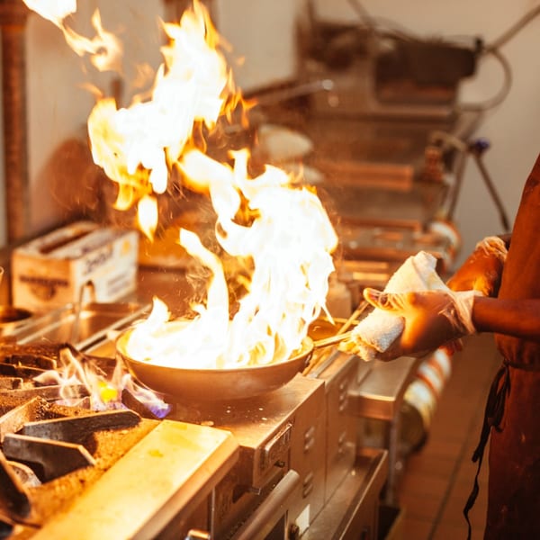 Chef flambéing food in a pan with large flames in a professional kitchen at Passage To India.
