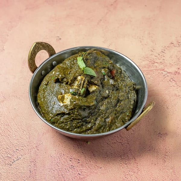 A metal bowl of vibrant green Palak Paneer, garnished with a fresh mint leaf, on a pink surface.